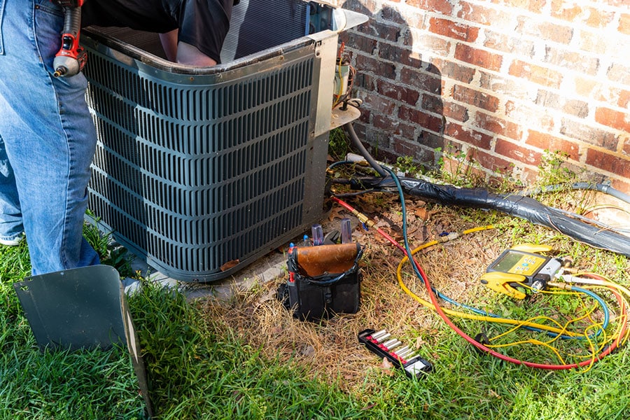 A technician performs AC repair on an outdoor unit beside a brick wall in Warren, MI, with tools, wires, and a tool bag scattered on the grass. Only the technician's legs and tool belt are visible.