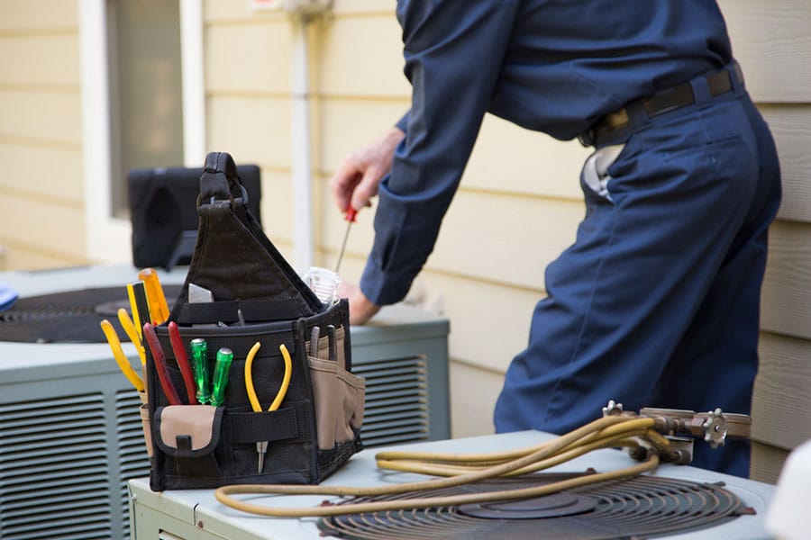 A technician in a blue uniform works on an outdoor HVAC unit next to a house in Warren MI, with a tool bag holding pliers, screwdrivers, and other AC repair tools placed on top of the equipment.