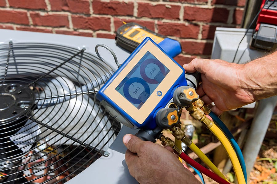 A person uses a digital manifold gauge to check the pressure on an outdoor air conditioning unit near a brick wall in Warren MI, with colored hoses connected—showcasing precision AC repair.