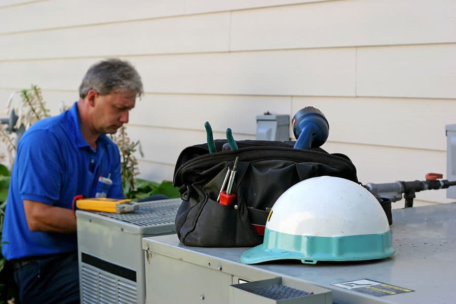 A technician in a blue shirt works on outdoor air conditioning equipment next to a New Blatimore, MI home. In the foreground, a tool bag, various tools, and a white safety helmet rest on the AC repair unit.