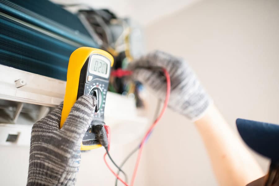 A person wearing gloves uses a digital multimeter to check an air conditioning unit during an AC repair in Sterling Heights MI, holding the device and connecting its probes to the equipment.