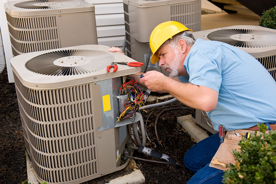 A technician wearing a yellow hard hat repairs an outdoor air conditioning unit, using a screwdriver to work on exposed wires—potentially addressing air conditioner problems like an AC water leak—with tools in his belt and multiple AC units nearby.