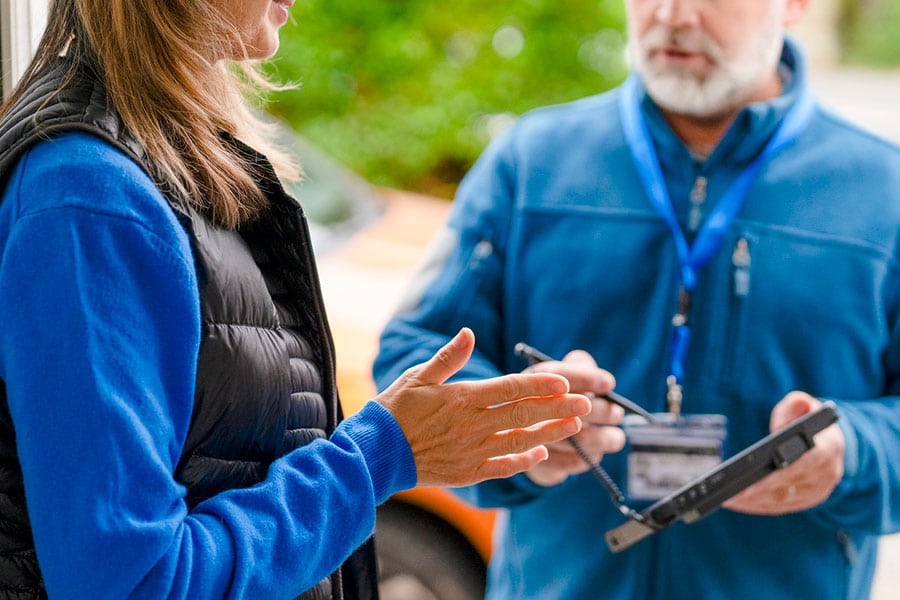 A woman gestures while talking to a man in a blue jacket holding an electronic tablet and stylus, possibly filling out a form or survey outdoors. They could be discussing air conditioning options for Michigan homeowners or addressing common FAQs about home comfort solutions.